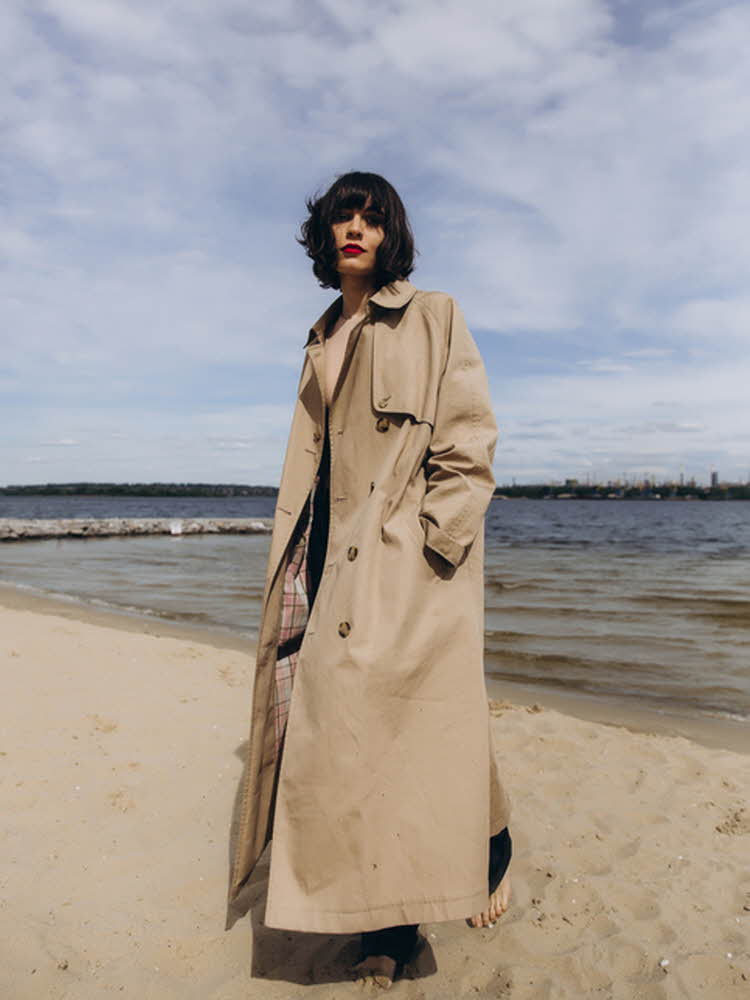 Dame med mørkt, kort hår poserer på strand med stor, beige trenchcoat
