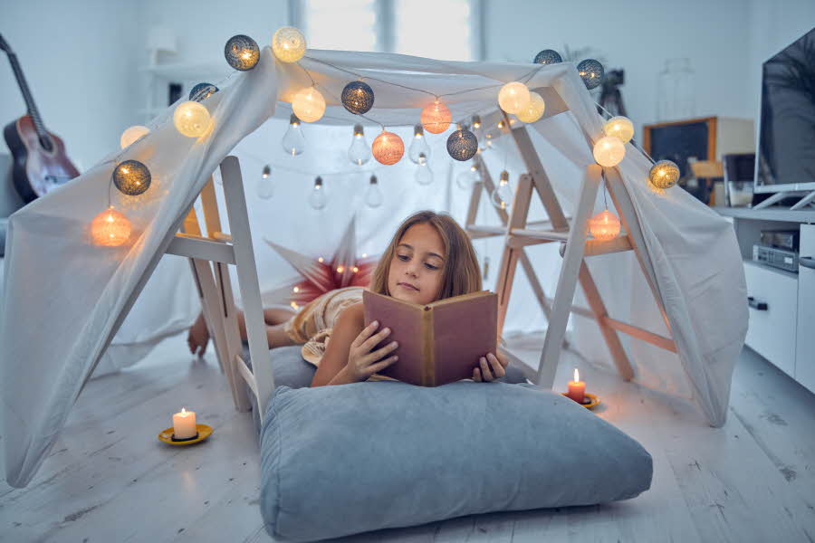Jente som leser bok på gulvet i sitt hjemmelagde telt i stua
