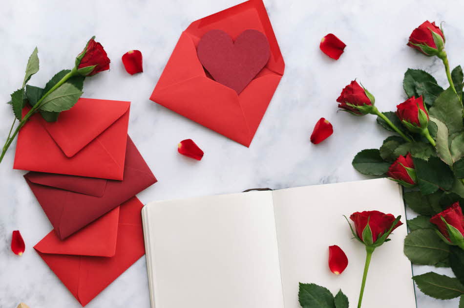 Åpen bok på bord med røde konvolutter og røde roser rundt