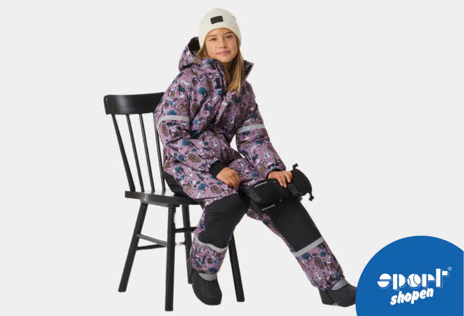 Jente sitter på en stol iført mønstret lilla vinterdress med refleksdetaljer og vinterstøvler.