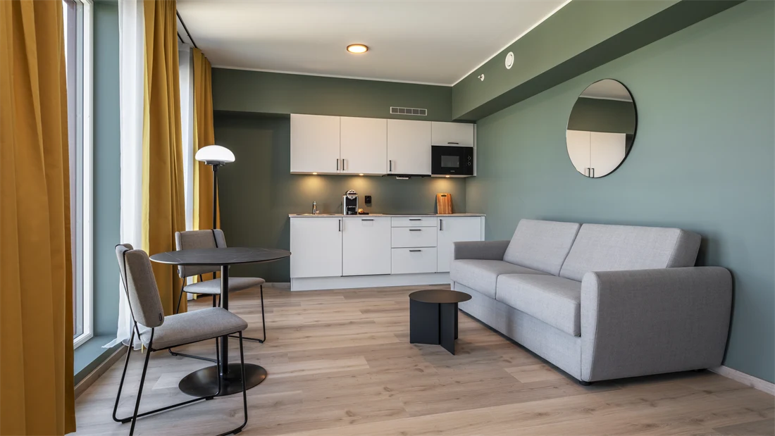 Business rom med kjøkken krok, grå sofa, spiseplass, grønne vegger, gule gardiner og parkett.
