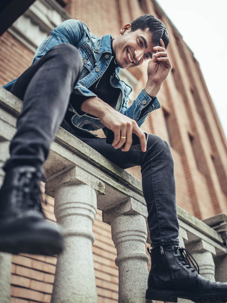 Mann med denimjakke, grå bukse og sorte boots sitter på gjerde sett nedenfra