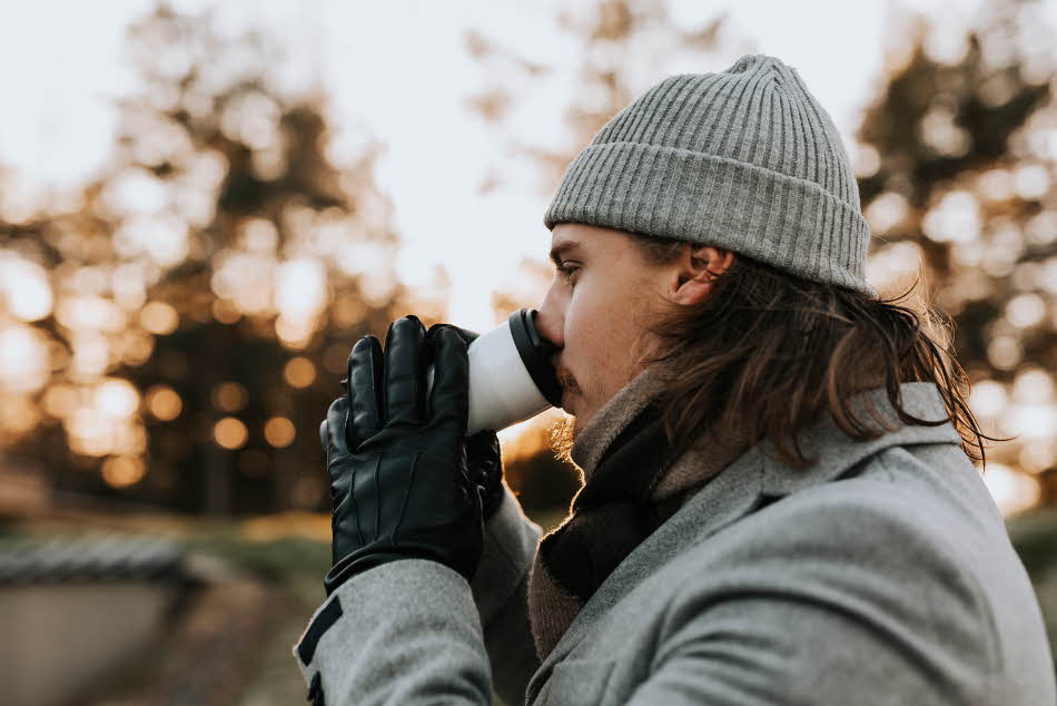 Nærbilde av mann med grå lue og sorte skinnhansker som drikker kaffe sett fra siden