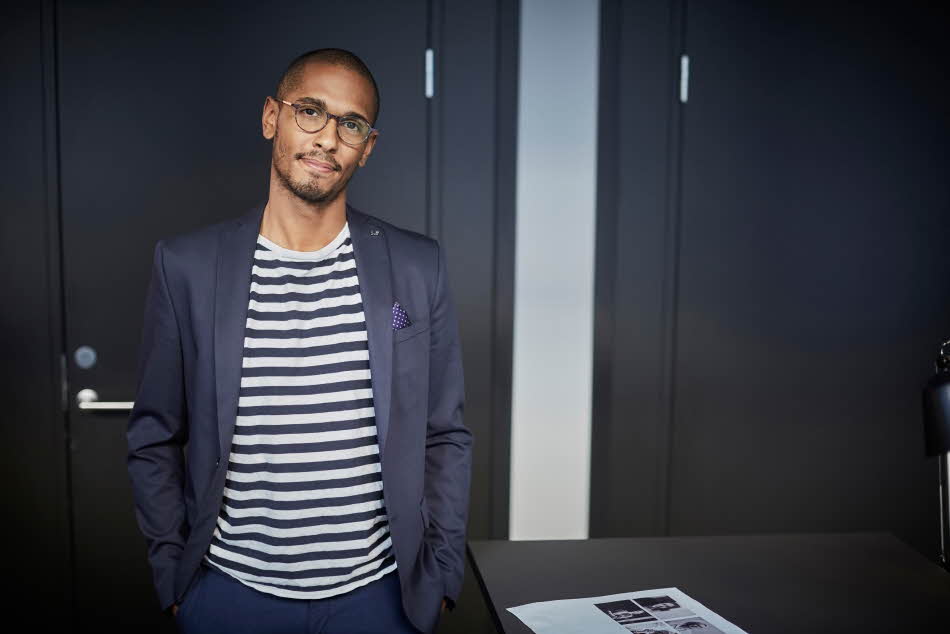 Mann med blå dress, hvitstripete t-skjorte og briller poserer til kamera inne på kontor