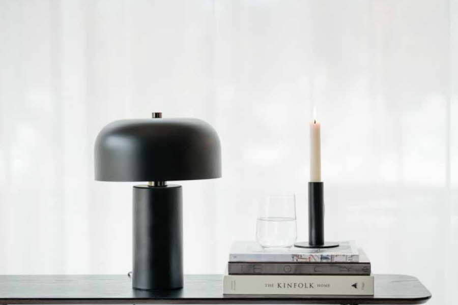 En svart lampe ved siden av tre bøker, en lysestake og et glass med vann på en svart hylle