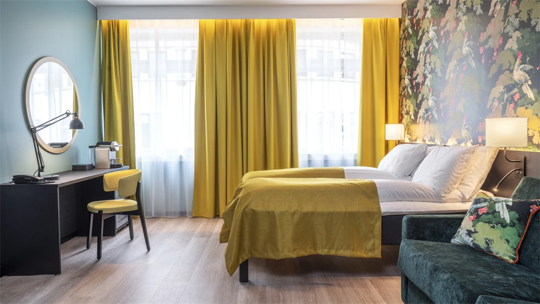 Turkis vegg, vegghent speil, skrivebord og gul stol. Store vinduer med gule gardiner. Fargerik tapet, seng med gult sengeteppe og grønn sofa i familie rom på Thon Hotel Bristol Bergen