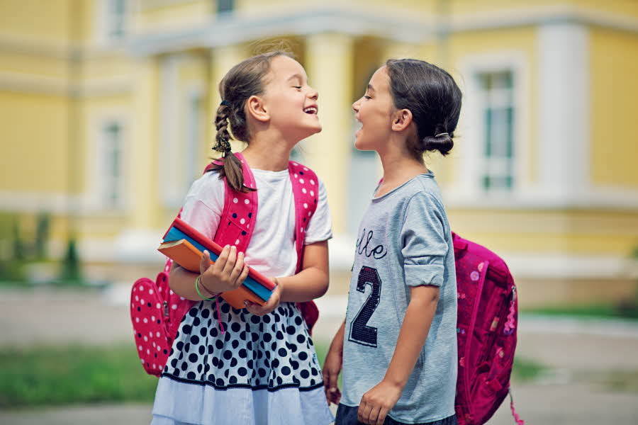To jenter med skolesekk og bøker som ser på hverandre og smiler
