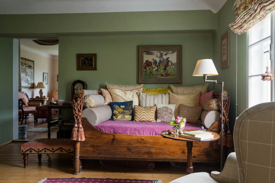 Grønn stue med puff, stålampe, lenestol og en gammel sengebenk full av puter.