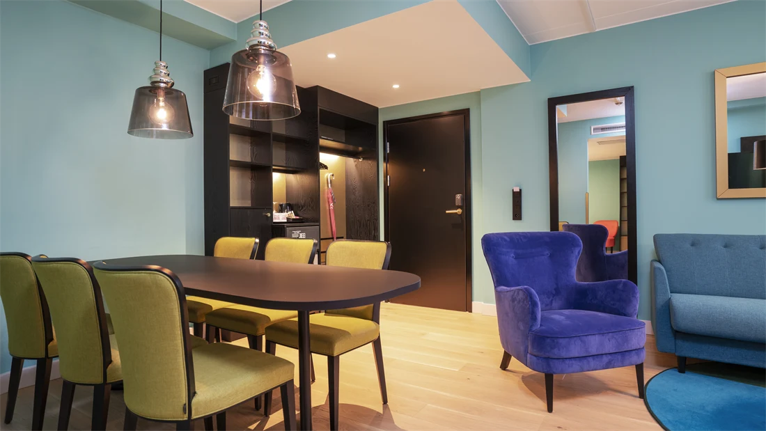Turkis tapet, spisebord med gule stoler og mørkt bord, stort speil, sittegruppe med turkis siofa og blå stol, i junior suite på Thon Hotel Storo i Oslo