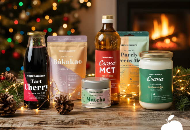 Supernature-produkter som kakao, matcha, kokosolje og MCT-olje plassert på bord med julelys og peis i bakgrunnen.