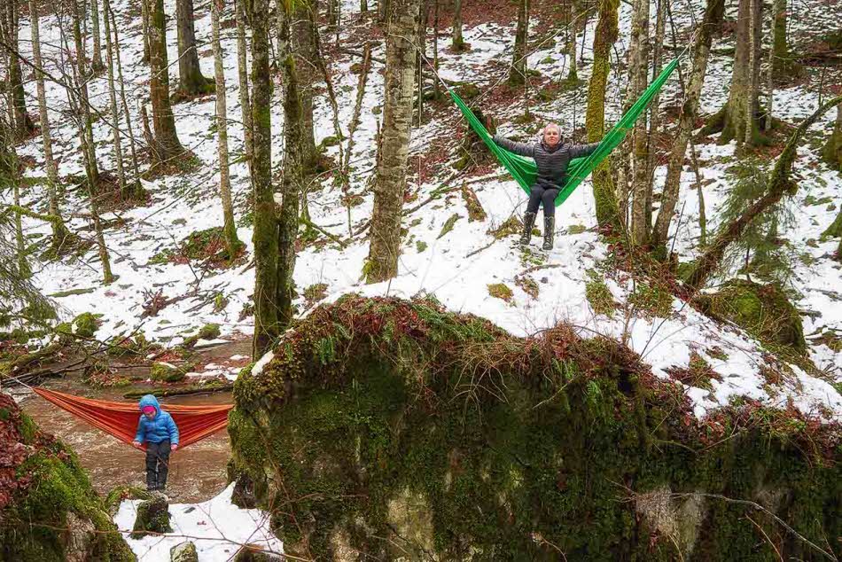 To personer i hver sin hengekøye mellom trær i delvis snødekket skog