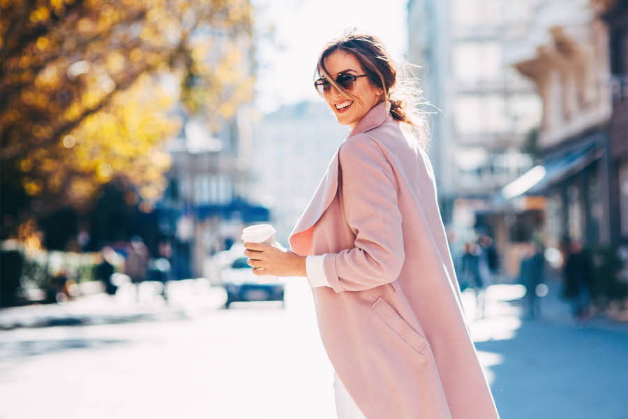 Dame smiler iført lys rosa kåpe og solbriller på gata med kaffe i hånda.