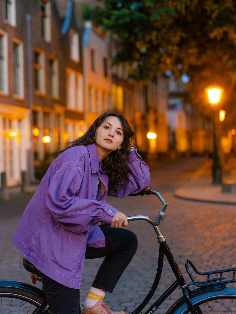 Dame med mørk lilla olajakke og sort bukse sitter på sykkel med hus og trær i bakgrunnen