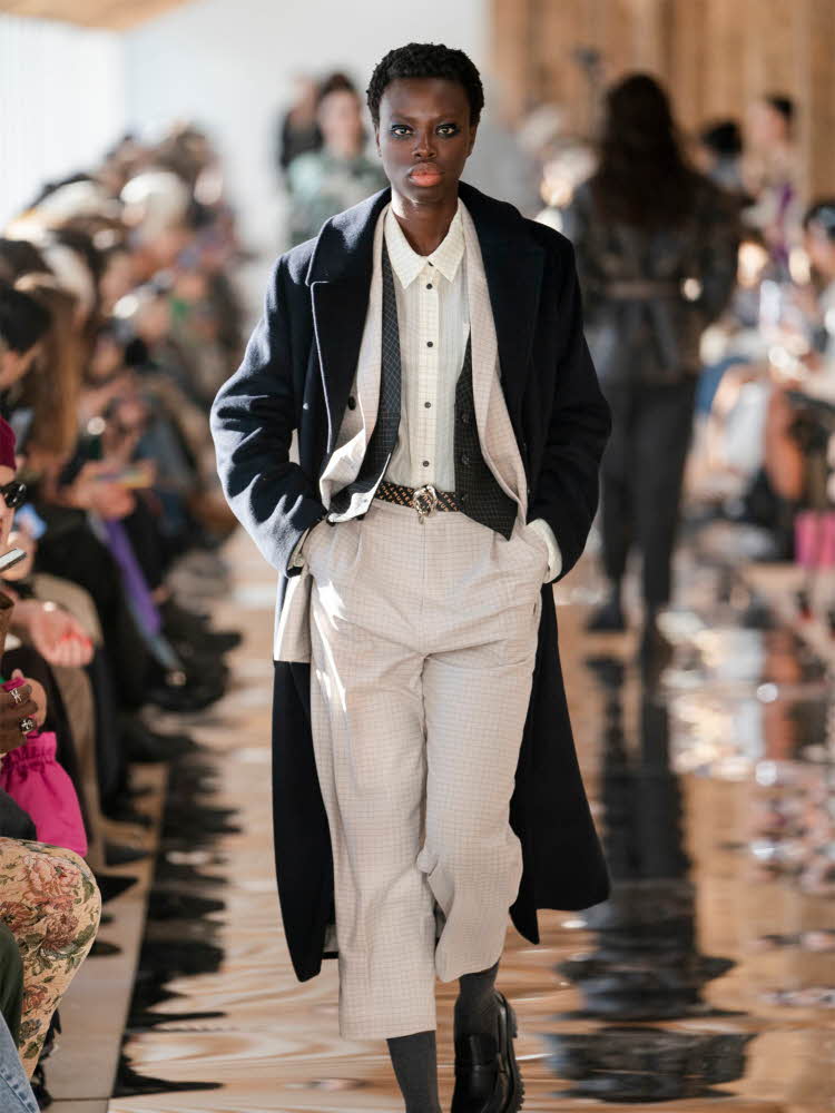 Damemodel på catwalk kledd i hvit skjorte, blazer, blå lang kåpe, hvite bukser og sorte sko.