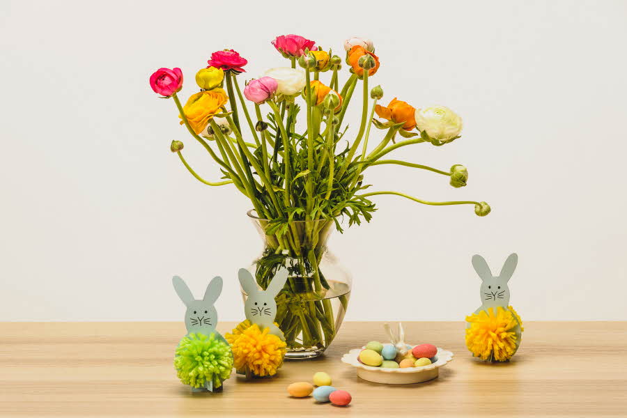 To grå kaniner med dusk laget av grønt og gult garn ved siden av en stor vase med blomster og sjokoladeegg på ett bord
