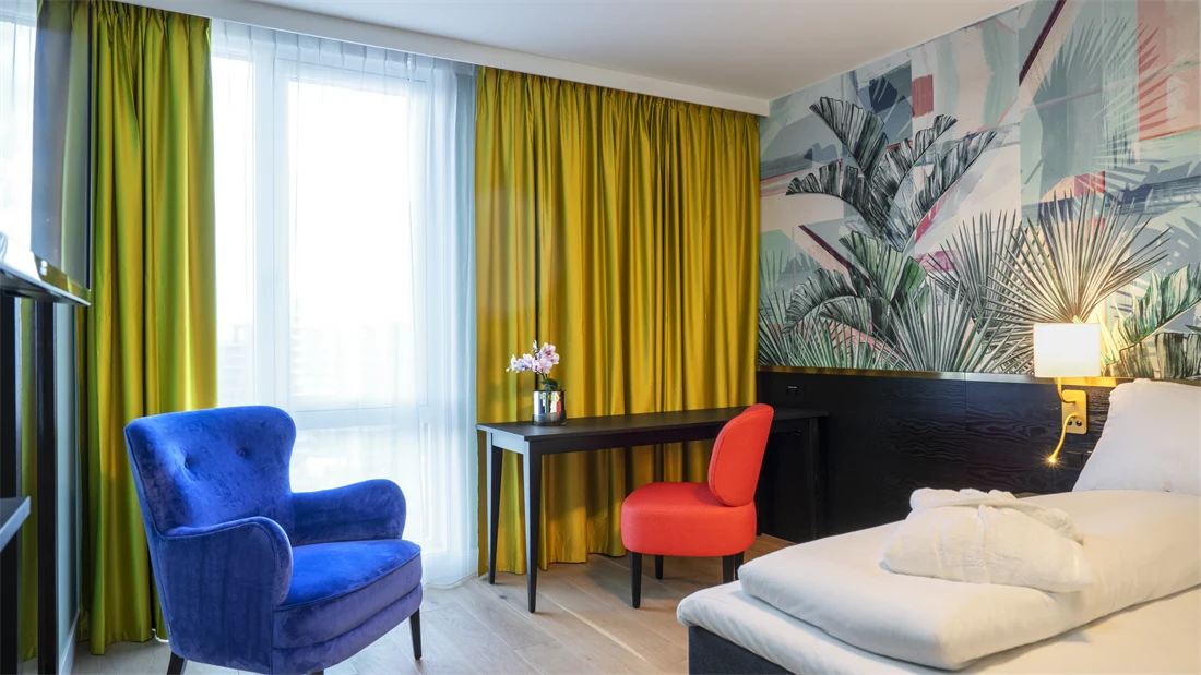 Del av dobbeltseng, arbeidspult og stor blå lenestol, store lyse vinduer og gule gardiner på dobbeltrom på Thon Hotel Storo i Oslo