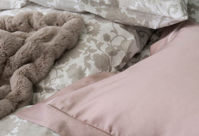 Seng med blomstrete sengetøy, rosa putevar og beige pelspledd.