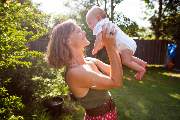 Kvinne smiler og løfter opp baby i hage på sommeren