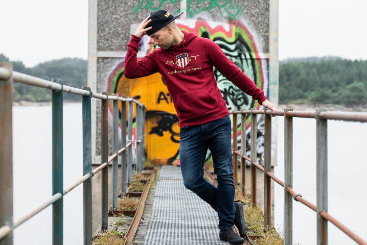 mann som står på en bro med et tårn bak seg. På veggen bak er det tagget. Han har på seg rød hettegenser, mørkt jeans og en caps. Han lener seg bort fra gelenderet og tar på capsen sin.