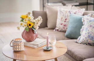 En stue med en sofa som har flere pynteputer, og et stuebord med levende lys og en vase med blomster.