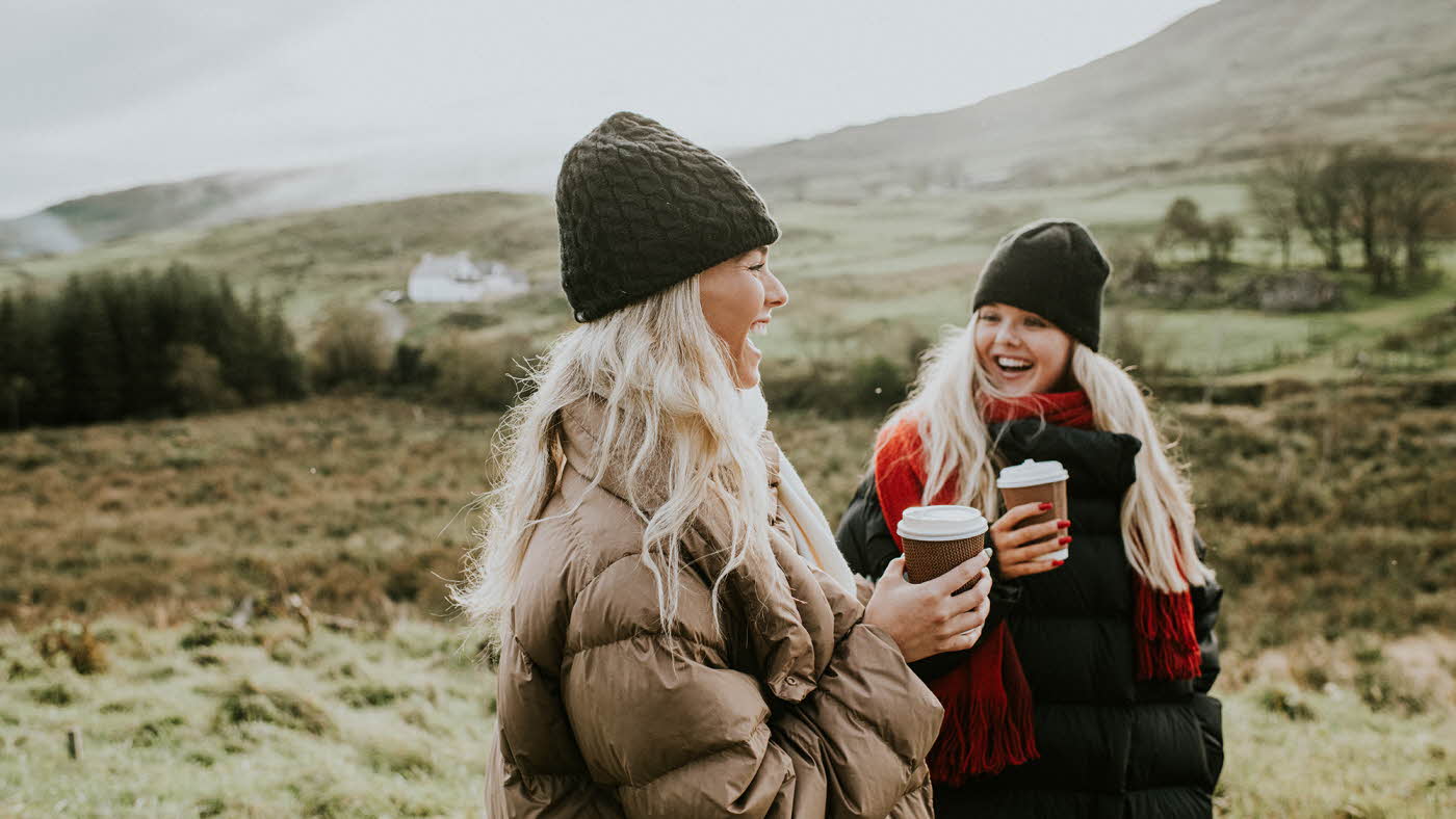 To jenter i vinterjakker går og prater med kaffe i hånda 