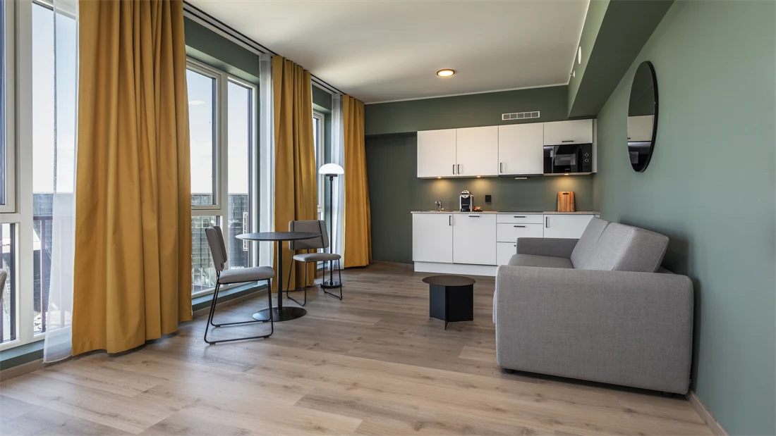 Business rom med kjøkken krok, grå sofa, spiseplass, grønne vegger, gule gardiner og parkett.