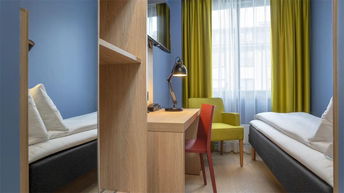 Speil, garderobe, skrivebord, TV, blå vegger, gul stol, gule gardiner og enkeltseng på lite enkeltrom på Thon Hotel Astoria