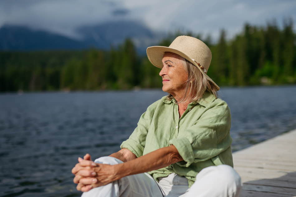 Eldre dame med pastellgrønn skjorte og stråhatt sitter på bryggekant og ser utover vannet