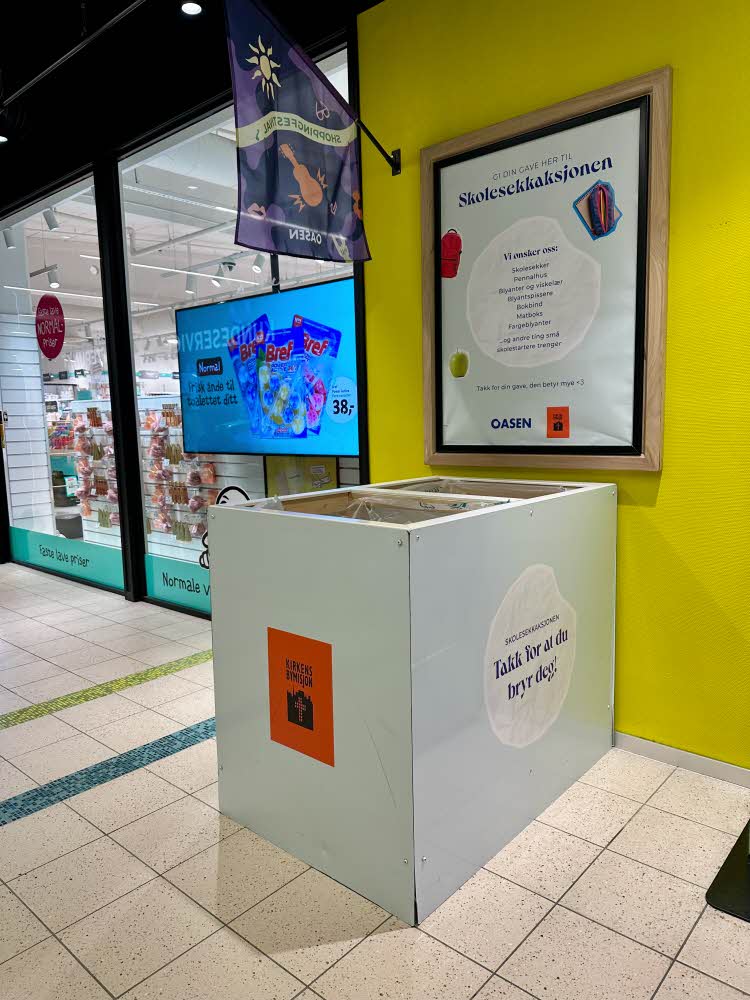 en stor blå pappeske hvor man kan donere skolesekker står opp mot en gul vegg med en plakat med informasjon om aksjonen over.