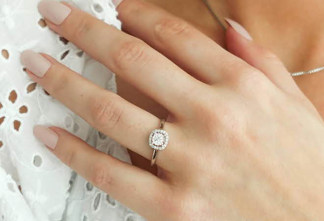 Hånd med elegant diamantring i halo-design på hvit blondebakgrunn.