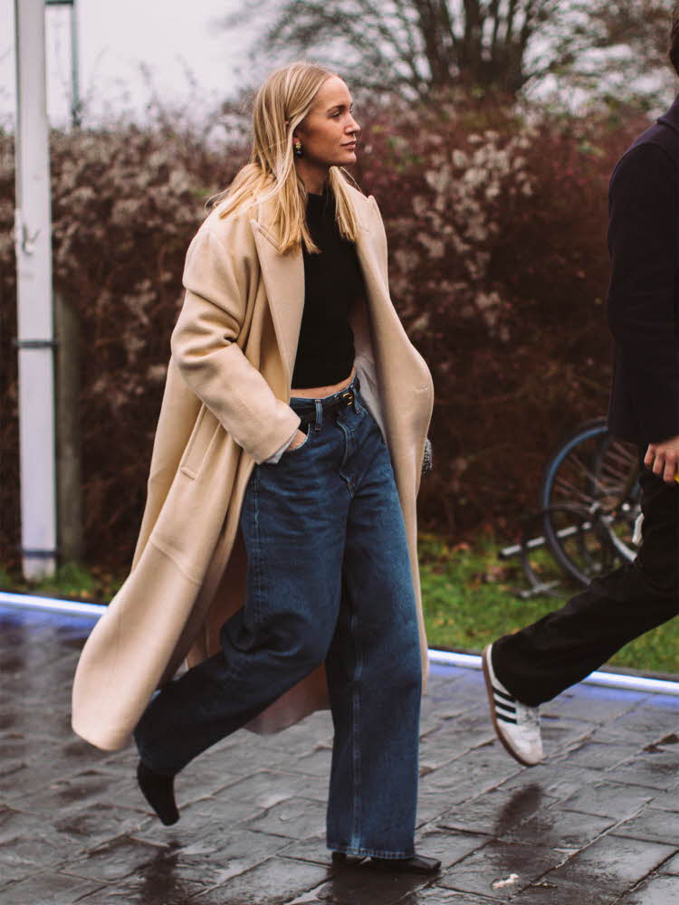 Gatefoto av dame med langt hår, lang beige kåpe, sort topp, jeans og sorte boots.