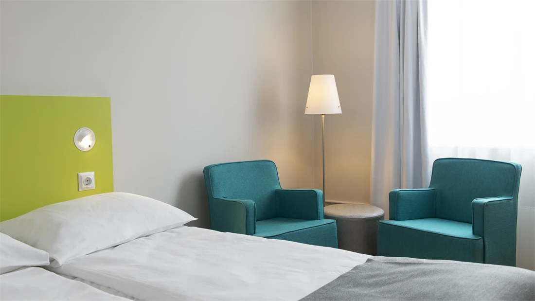 Nærbilde av Standard Room Twin på Thon Hotel Trondheim. Hvit vegg, limegrønt sengegavl, to oppredde enkeltsenger, to turkise lenestoler, lampe og et lite rundt bord.