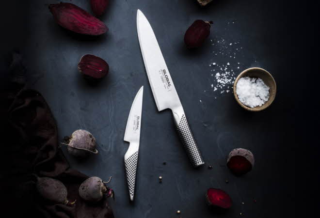 To kniver fra Global som ligger på et bord med grønnsaker rundt