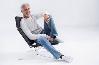 En mann i grå genser og jeans sitter i en stol og smiler mot kamera.